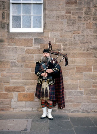 Scottish bagpiper in traditional kilt and uniform stands outdoors against a stone wall playing bagpipes, window and cobblestone visible.