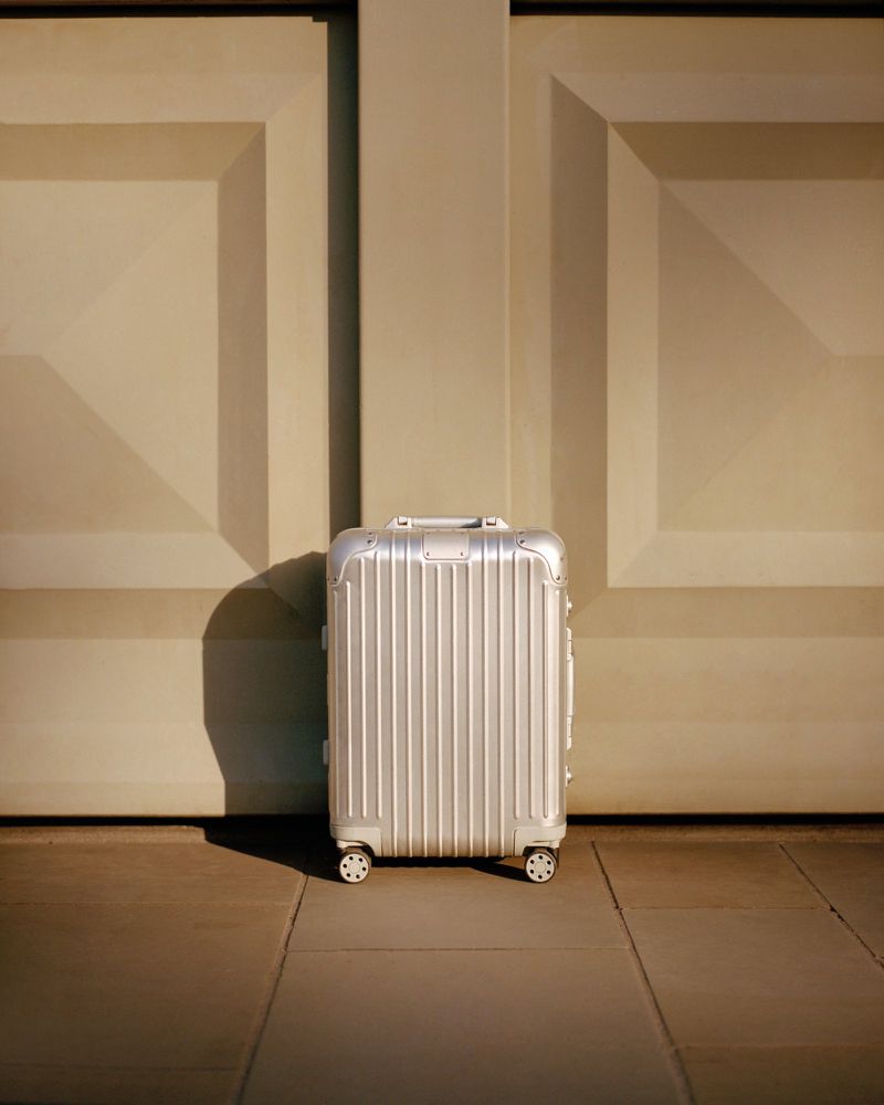 Silver suitcase stands on a brown tiled surface in front of a cream coloured wall with square shapes and subtle shadows.