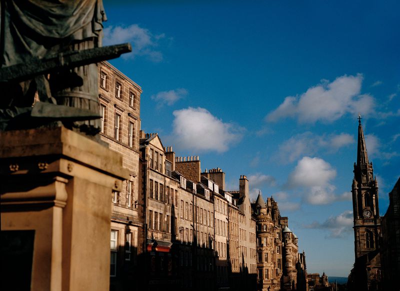 Urban street view in Edinburgh, Scotland, features aged stone buildings, church steeple, and blue sky with scattered clouds on a sunny day.