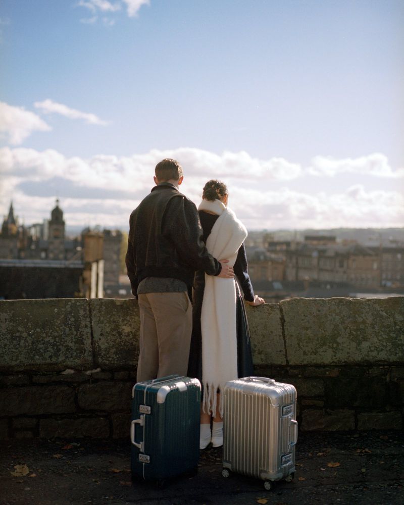 A couple stands on a stone wall overlooking a European cityscape with two suitcases, on a sunny day with blue sky and white clouds.