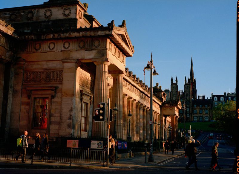 People walk on the sidewalk near a historic building with columns under a clear sky in the city, with cars on the road in the distance.