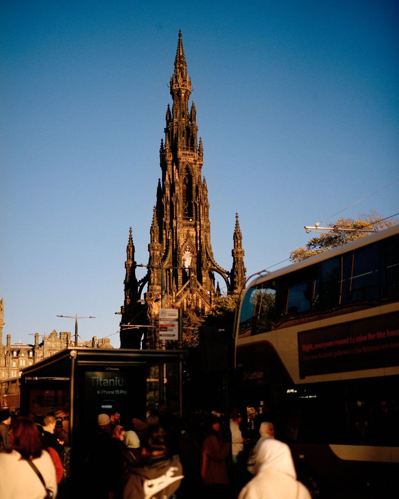 The Scott Monument in Edinburgh, Scotland, towers over a crowd and a passing double-decker bus under a bright blue sky in an urban setting.