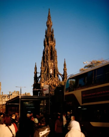 The Scott Monument in Edinburgh, Scotland, towers over a crowd and a passing double-decker bus under a bright blue sky in an urban setting.