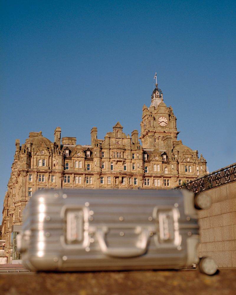 Suitcase in front of historic sandstone hotel building with clock tower on a sunny day under a clear blue sky.