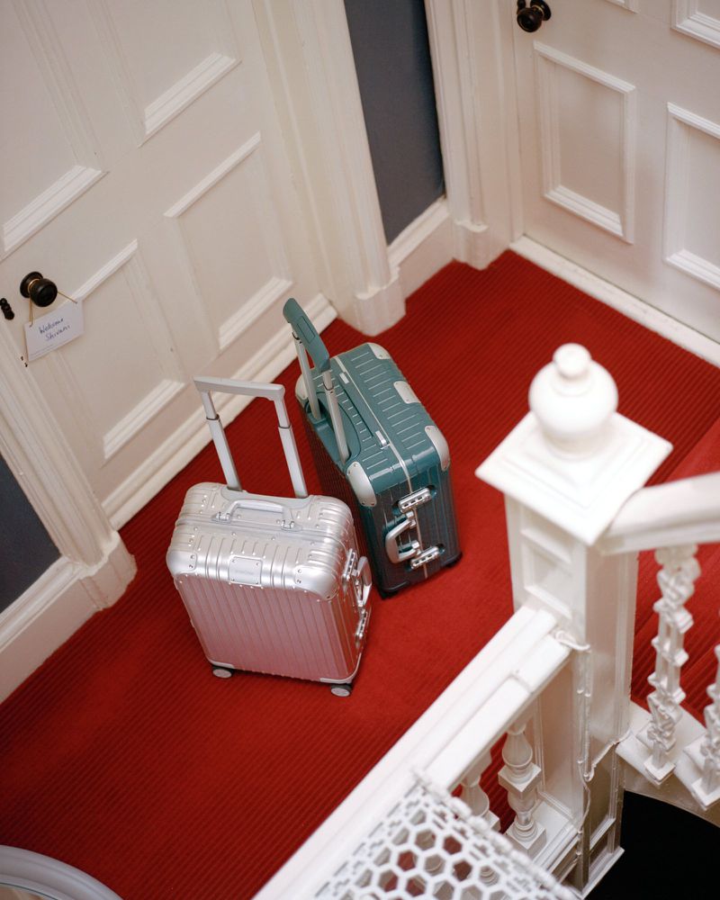 Two suitcases, a silver roller and a teal suitcase, rest on a red patterned carpet in a hallway with white doors and banisters.