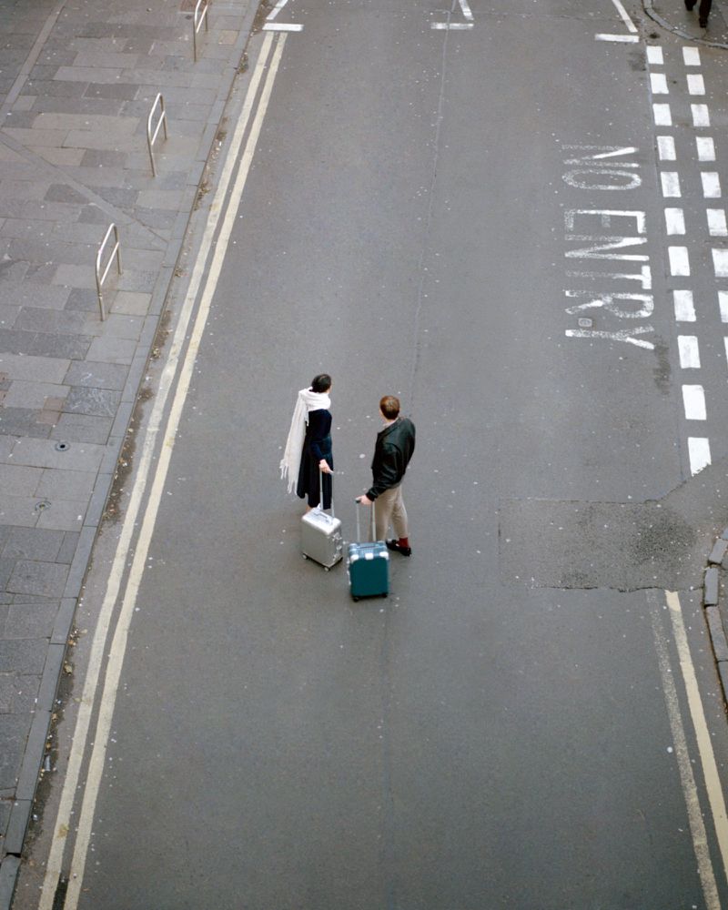 High angle view of a stylish couple standing on a city street with their suitcases, ready for a trip, under soft, natural light.