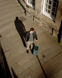 Man standing on stone pavement with a green suitcase, casting a long shadow next to historic building steps.