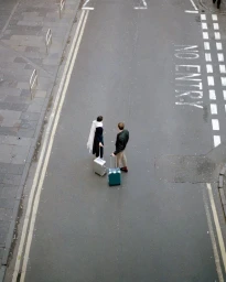 High-angle view of two people walking on a city street pulling wheeled suitcases next to road markings.