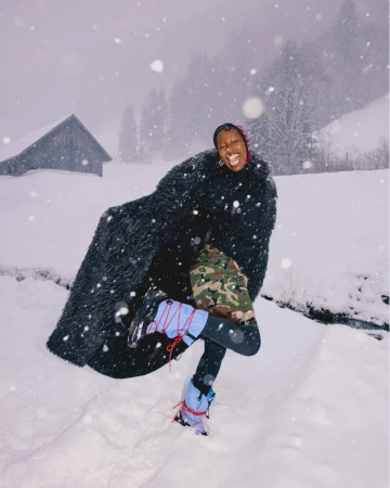 A happy Black woman stands on snow-covered ground in winter, wearing a long fur coat, colorful pants and boots, with snowy mountains visible.