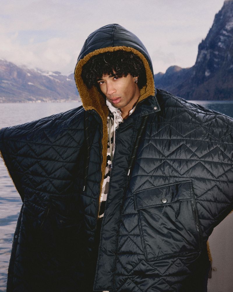 Adult male poses outdoors in a quilted poncho with a fur-lined hood, showcasing a unique fashion statement against a natural backdrop.