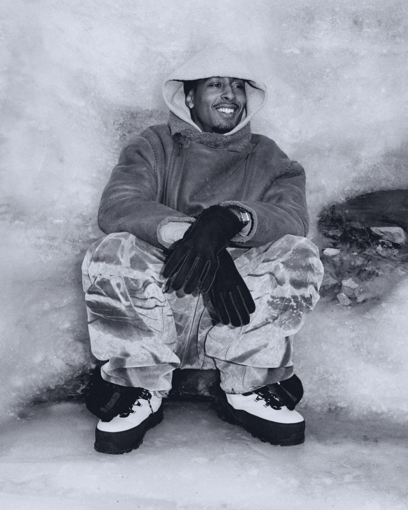 Man wearing hooded coat, gloves, and snow boots sitting in a snowy or icy outdoor environment, smiling, looking to the side in a black and white photo.
