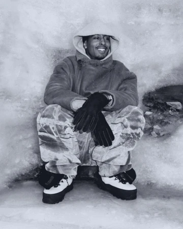 Man wearing hooded coat, gloves, and snow boots sitting in a snowy or icy outdoor environment, smiling, looking to the side in a black and white photo.