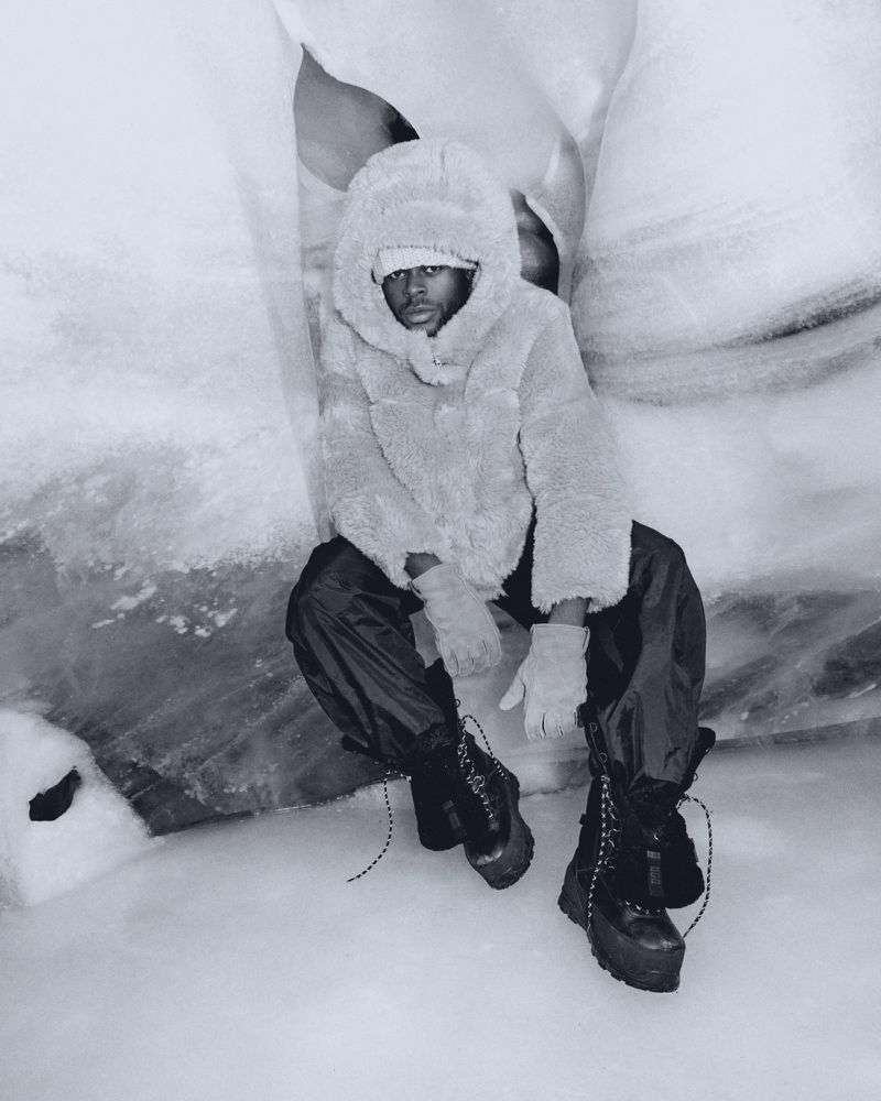 Man in furry hooded jacket, black pants, and platform boots sits in snow cave for fashion editorial photography in a stark monochromatic palette.