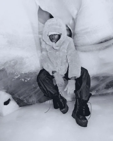 Man in furry hooded jacket, black pants, and platform boots sits in snow cave for fashion editorial photography in a stark monochromatic palette.