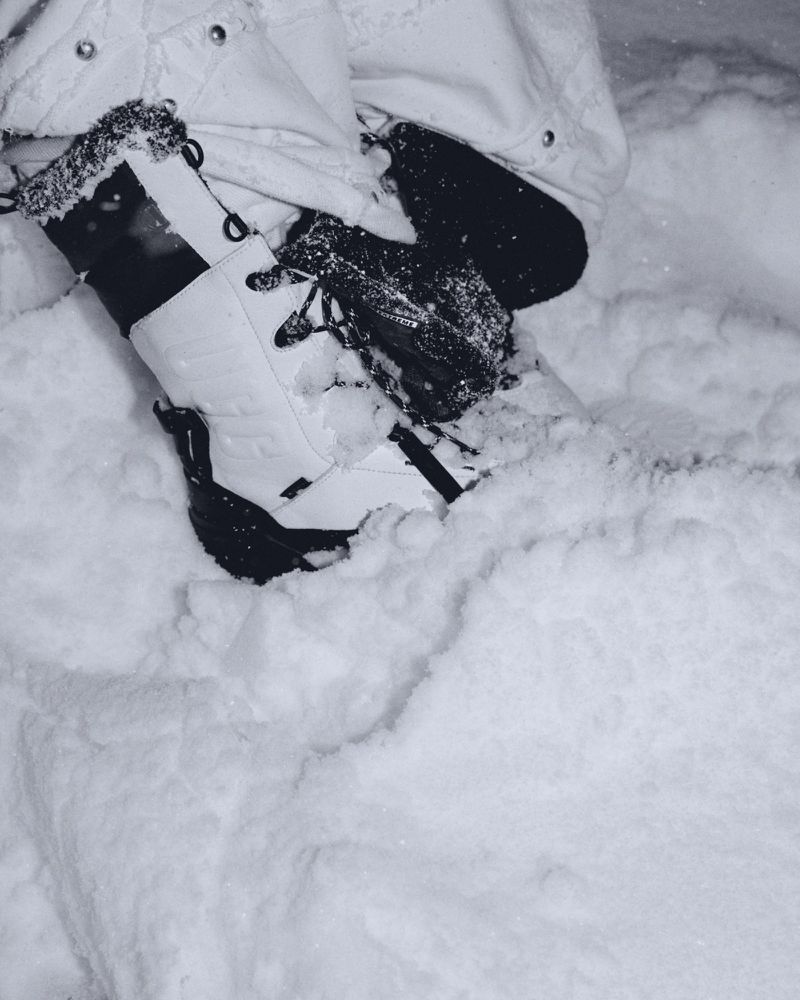 Close-up on person wearing white snow boots and black gloves, surrounded by fresh snow. Focus on footwear, apparel, and winter setting.