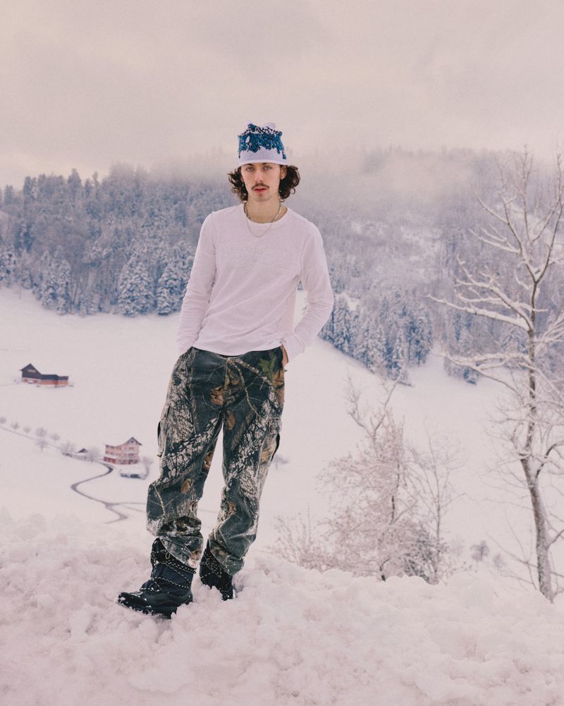 Young adult poses in the snow with winter forest in the background. He is wearing a hat and camo pants on a winter day.