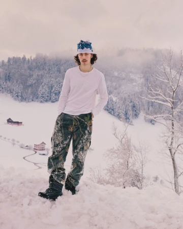 Young adult poses in the snow with winter forest in the background. He is wearing a hat and camo pants on a winter day.
