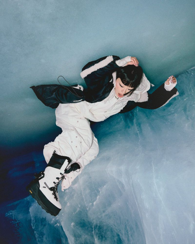 Overhead shot of woman in a black-and-white jacket and white snow pants and boots laying on a textured light blue surface.