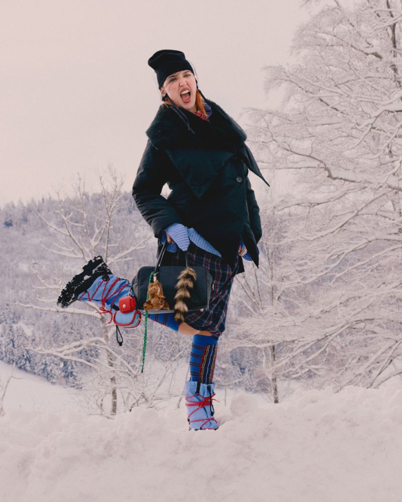 Fashionable woman in winter coat and boots poses playfully in snowy outdoor scene with trees, holding a stylish purse.
