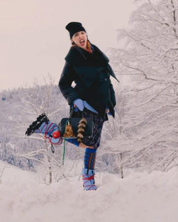 Fashionable woman in winter coat and boots poses playfully in snowy outdoor scene with trees, holding a stylish purse.