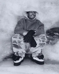 Man wearing a hooded winter jacket, gloves, and snow boots crouching against an icy snow-covered background.