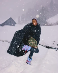 Smiling model wearing a black furry coat and blue winter boots outdoors in heavy snow