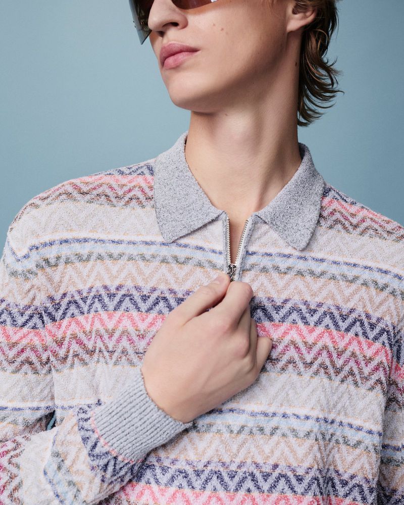 Close-up of a young adult man in a patterned knit polo shirt with a zipper, wearing sunglasses, set against a light blue backdrop.