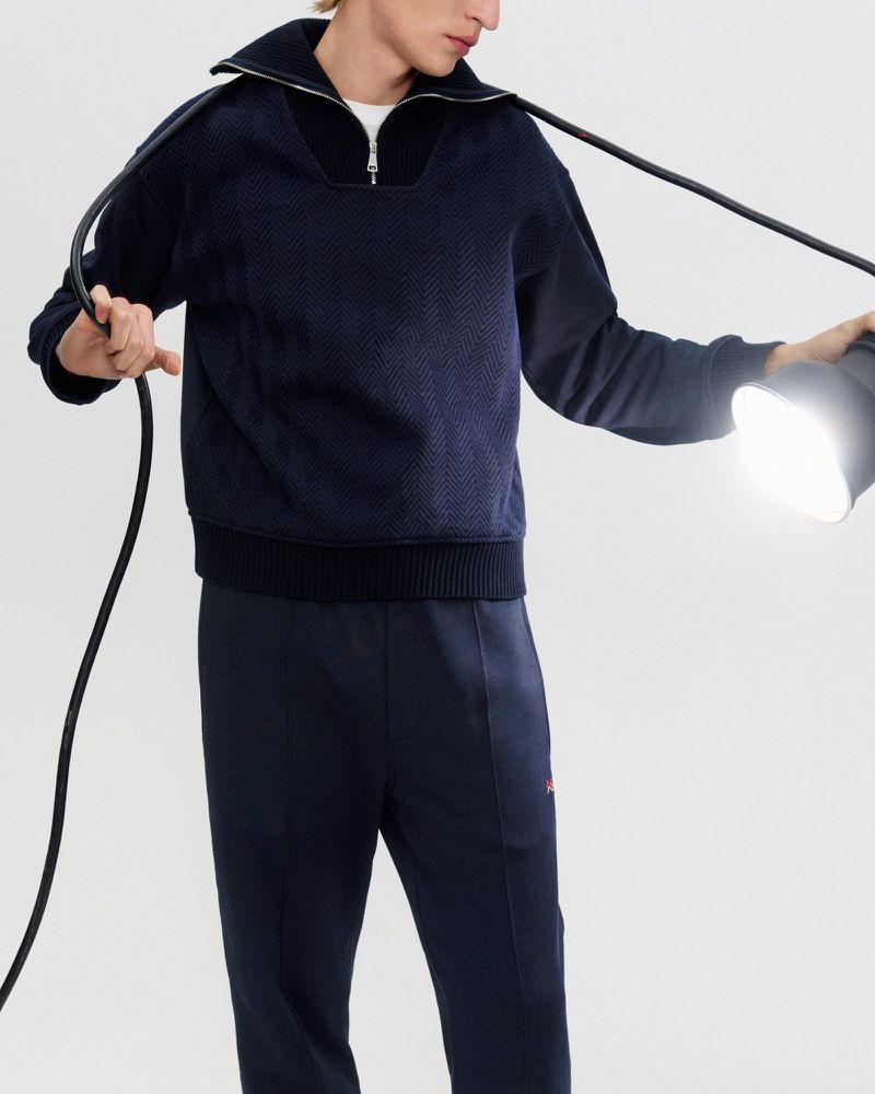 An adult man wearing a textured, dark-blue collared pullover and matching sweatpants stands under a bright spotlight indoors.
