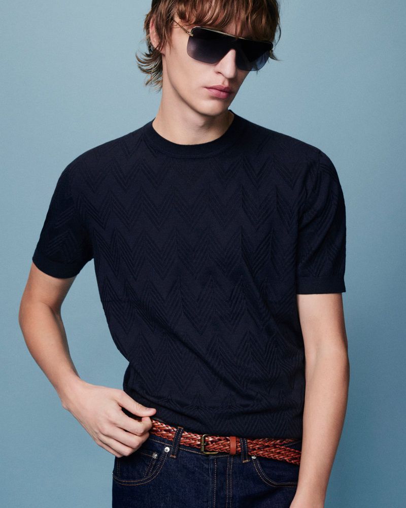 Man wearing sunglasses, dark textured t-shirt, and jeans with a brown braided belt stands in front of a blue studio background.
