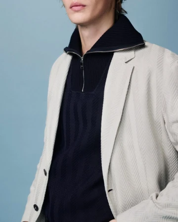 Man wearing a light grey blazer over a dark blue sweater with a quarter zipper, posing in front of a blue background.