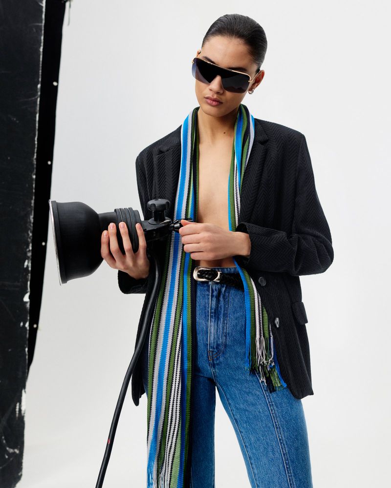 Adult female model wearing sunglasses, jacket, jeans, and scarf, holds a photo light in a photo studio setting. Fashion editorial shot.