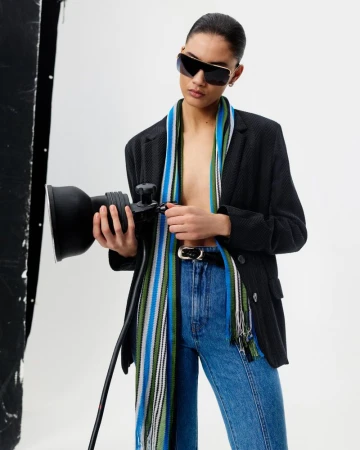 Adult female model wearing sunglasses, jacket, jeans, and scarf, holds a photo light in a photo studio setting. Fashion editorial shot.