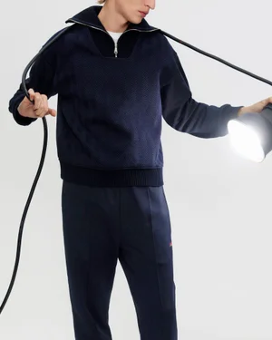 Model wearing navy zip-collar sweater holding a glowing studio lamp on white seamless background.