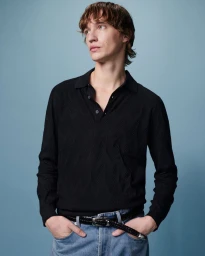 Model wearing a black knit polo shirt and blue denim jeans against a plain blue studio background