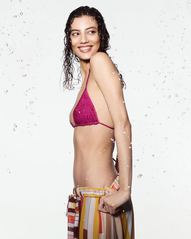 Smiling woman in pink bikini top and striped pants stands against a white background with water droplets, looking away from the camera.