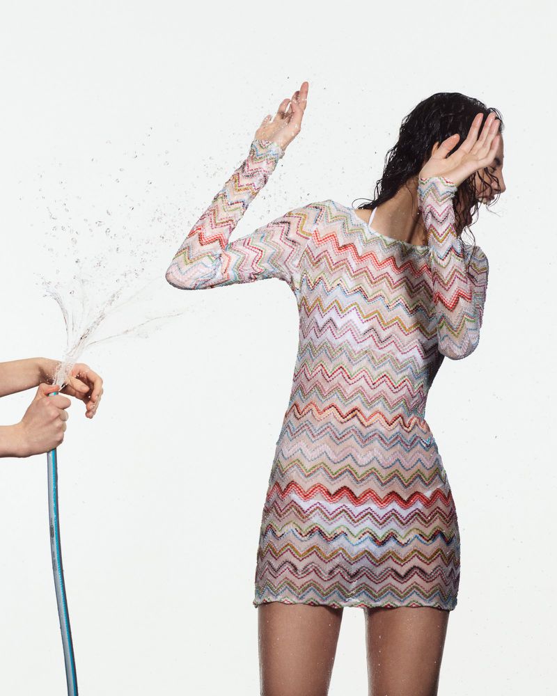 Woman in colorful zigzag mini-dress shields face from water hose on white background, creating a dynamic fashion shot.