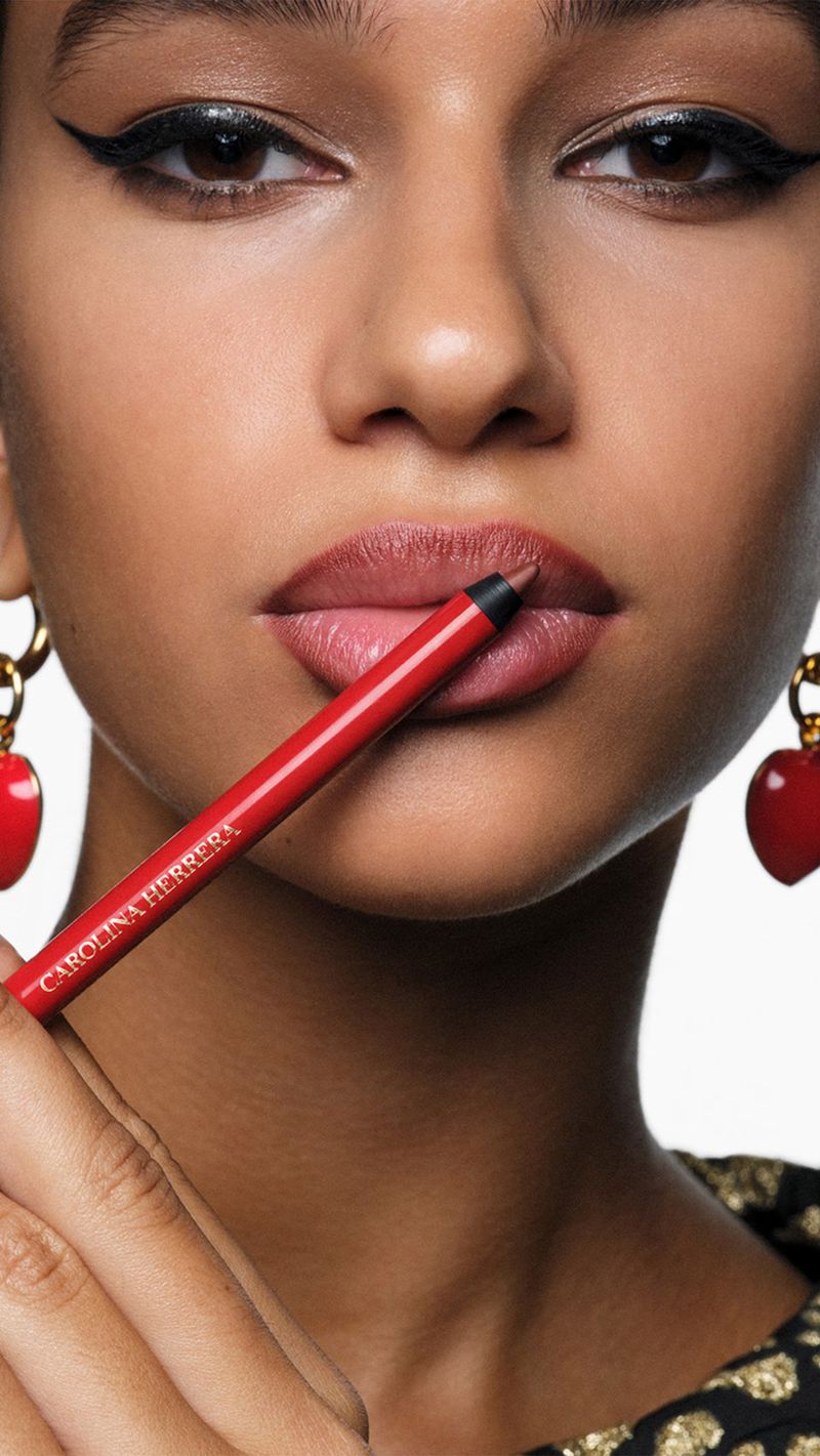 Close-up shot of a young woman with flawless skin applying red lip liner from Carolina Herrera, showcasing makeup artistry and cosmetic product.