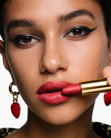 Close-up shot of a woman with red lipstick holding a lipstick tube. The focus is on the makeup and the gold earrings.