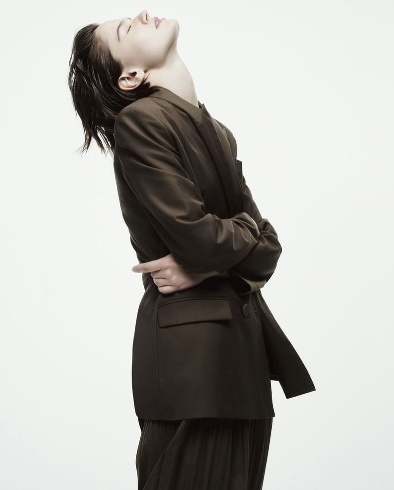 Woman in oversized olive blazer and pleated skirt poses in studio. Head tilted back, eyes closed, hands clasped behind her back.