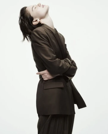 Woman in oversized olive blazer and pleated skirt poses in studio. Head tilted back, eyes closed, hands clasped behind her back.