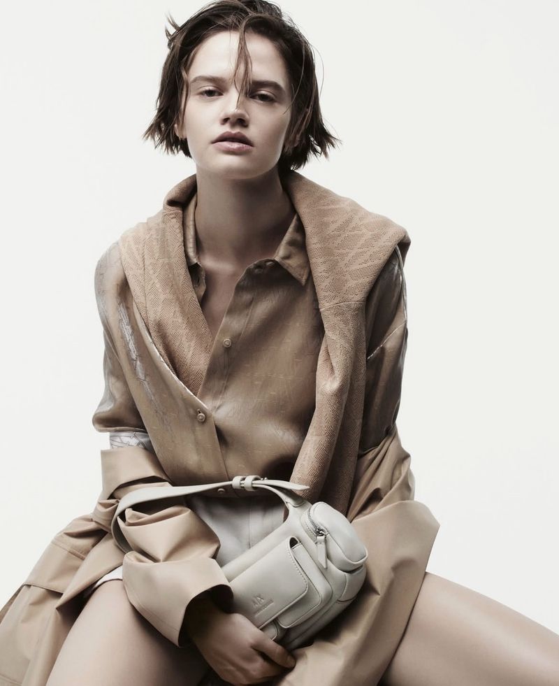A young adult fashion model poses while seated against a white background wearing a beige top, trench coat, and holding a white bag.