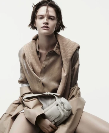 A young adult fashion model poses while seated against a white background wearing a beige top, trench coat, and holding a white bag.