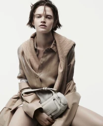 Model in camel shirt and monogram sweater holding a pale shoulder bag against white