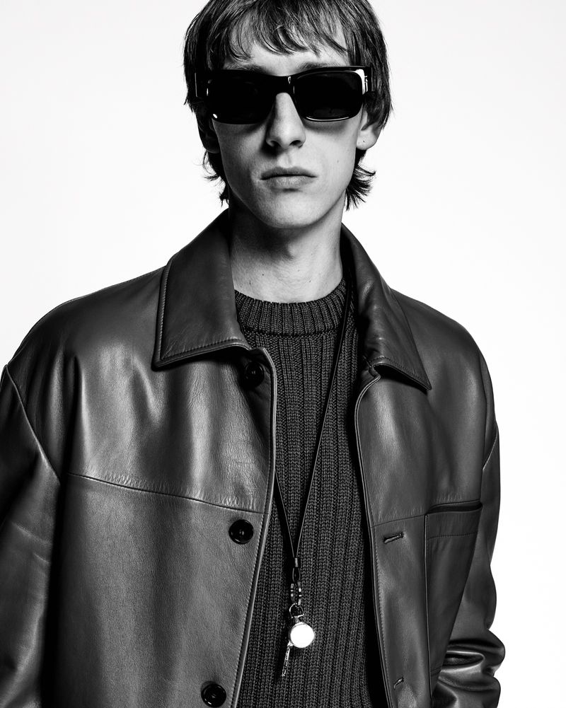 Monochrome portrait of a young adult male wearing sunglasses, leather jacket, and a sweater with a lanyard in a studio setting.