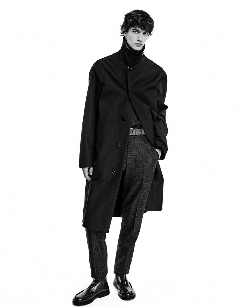Monochrome studio portrait of a male fashion model in a dark overcoat, turtleneck, patterned trousers, and shiny black Chelsea boots.