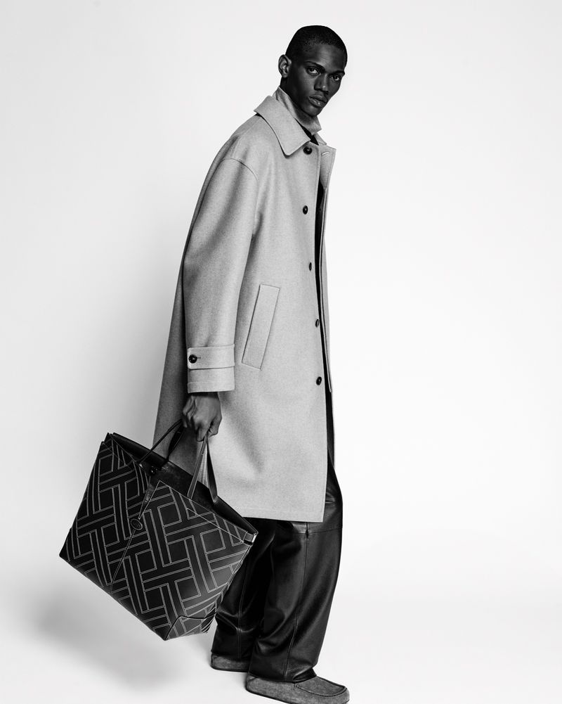 Monochrome fashion shot: a Black male model in a coat poses with a patterned tote bag against a bright white background in a studio.