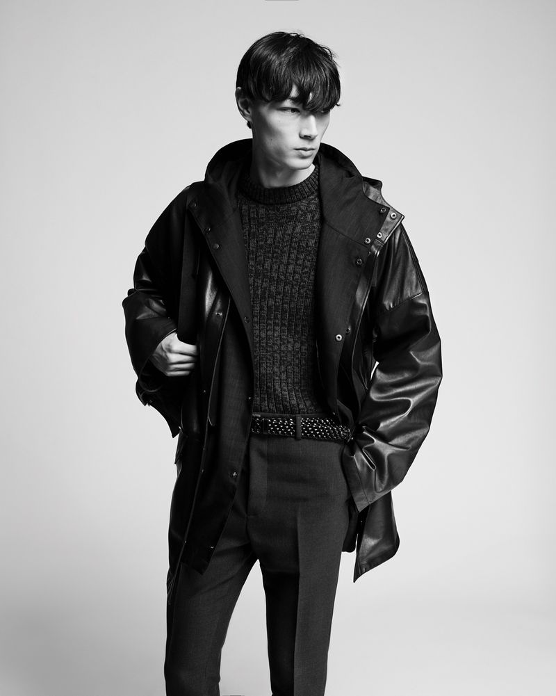 Monochrome fashion portrait of a young man in a dark hooded jacket, patterned sweater, and pants against a light backdrop.