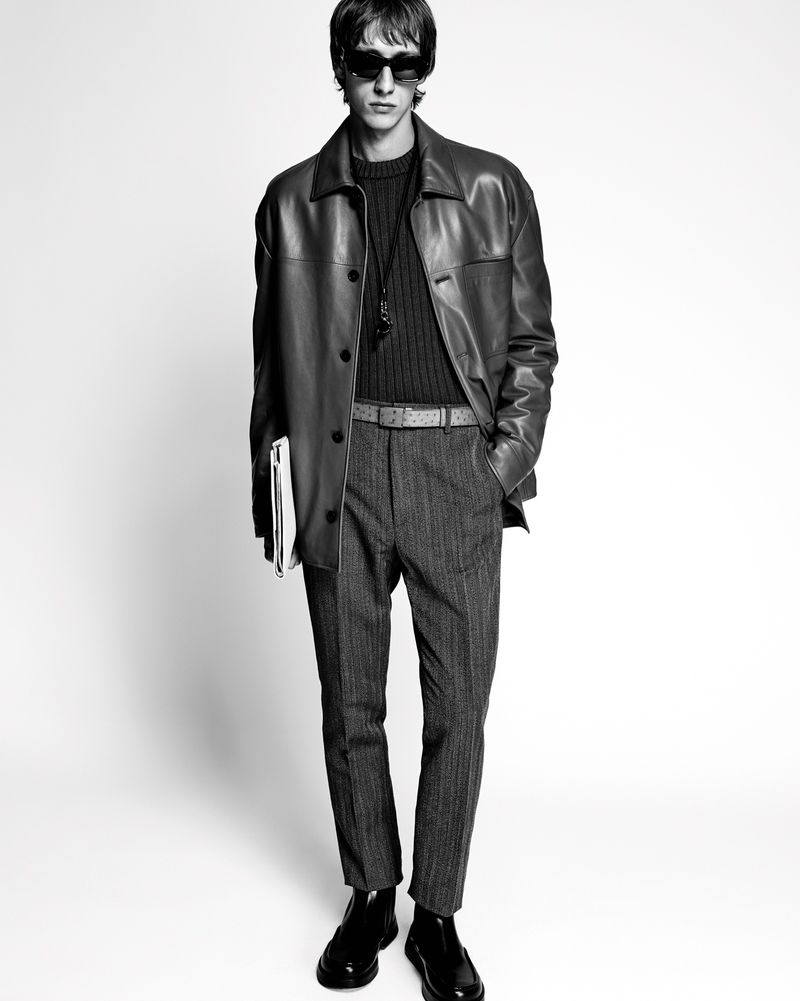 Monochrome shot of a man in sunglasses, a leather jacket, sweater, and patterned pants, holding documents, set against a bright background.