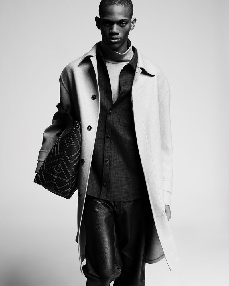 Black and white fashion shot of a dark-skinned male model wearing a coat, shirt, and trousers, and holding a patterned bag.