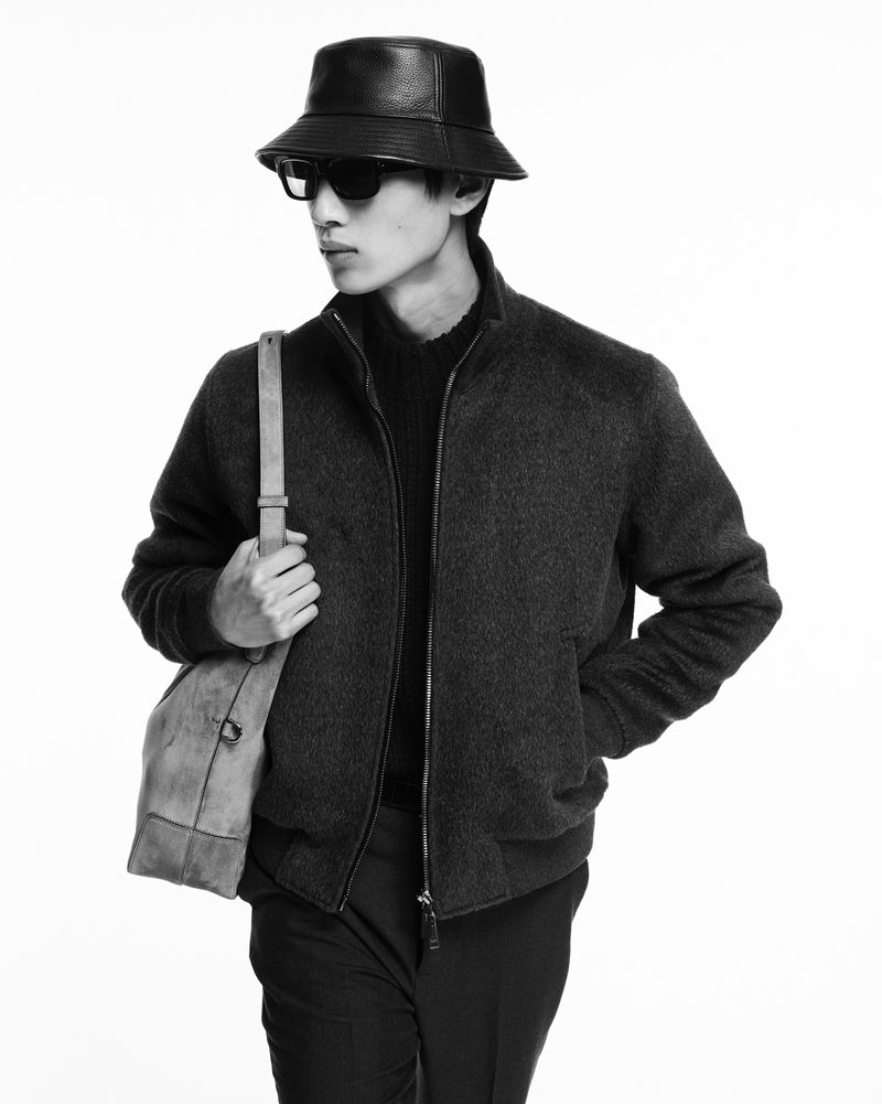 Black and white portrait of an Asian male fashion model wearing a bucket hat, jacket, sunglasses, and bag, posing in a photo studio.